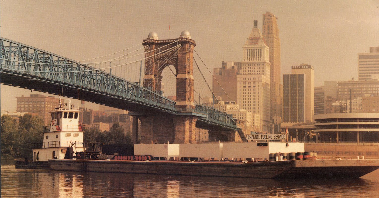 An archival photograph of a suspension bridge with a barge passing beneath